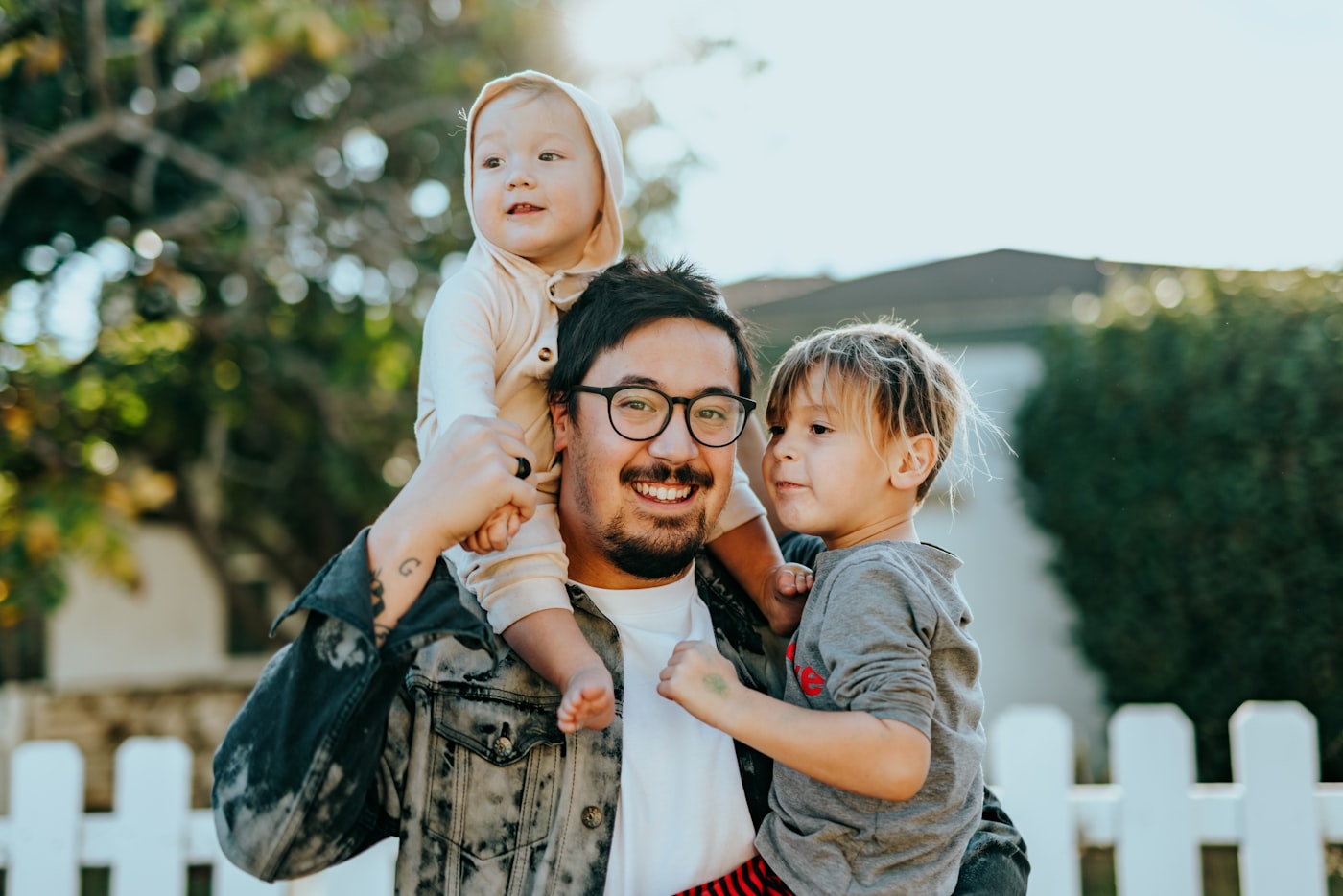 Pai sorrindo com dois filhos no colo em frente de casa